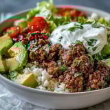 Colorful Low Carb Burrito Bowl with seasoned beef, cauliflower rice, melted cheddar, sour cream, cilantro, and lime wedges for squeezing.